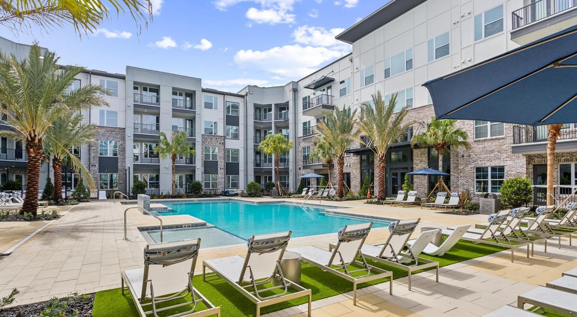 A swimming pool with lounge chairs, palm trees, and a multi-story building with balconies.