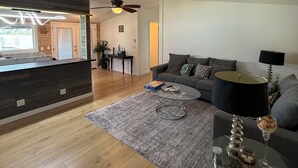 A modern living room with a wooden floor, a grey sofa, a coffee table, a glass table with a lamp, and a ceiling fan. - Lakefront 3BR/2BATH with Private Dock on 3/4 Acre in terrific Nocona with AC (Nocona)