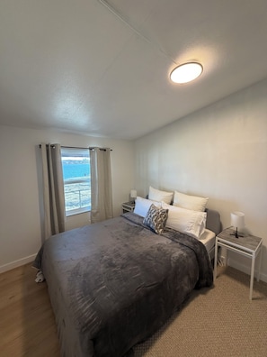 Un dormitorio con cama, mesitas de noche, ventana con cortinas y vistas al exterior.