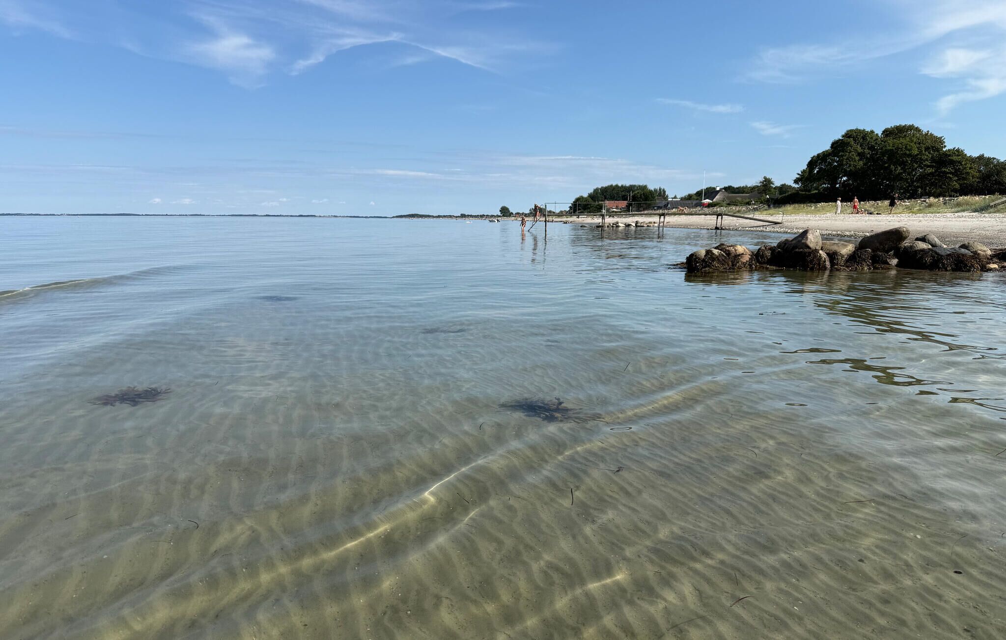 Ein sauberer Strand mit ruhigem Wasser, sandigem Ufer und einer entfernten Küstenlinie.