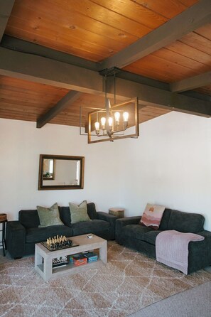 Ein gemütliches Wohnzimmer mit Holzboden, einem großen Spiegel, einem dunklen Sofa, einem Couchtisch mit Schachspiel und einem flauschigen Teppich.