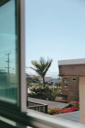 Blick aus einem Fenster mit Geländer, Palmen und einem Gebäude mit kleinem Fenster.