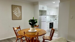 Uma sala de jantar com uma mesa de madeira redonda, quatro cadeiras de madeira, um vaso com planta e uma obra de arte com folha emoldurada na parede.