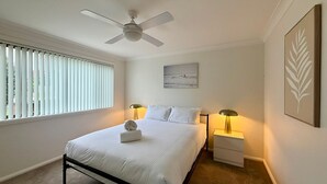 Quarto com uma cama, duas mesas de cabeceira com luminárias, um ventilador de teto, uma janela com persianas e um quadro na parede.