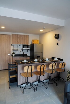 A modern kitchen with a central island, bar stools, a microwave, a refrigerator, and a wall-mounted clock.