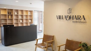 Aula, jossa on musta vastaanottotiski, puiset tuolit ja seinä, jossa lukee teksti: »VILLADHARA BARRIO-CREATIVO».