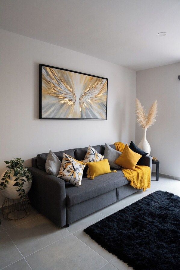 A modern living room with a grey sofa, yellow and grey cushions, a black rug, a large abstract painting, and a tall vase with pampas grass.