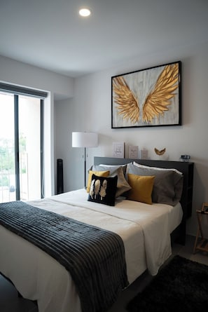A modern bedroom with a large bed, a view through a sliding door, a wall-mounted lamp, and a framed artwork of golden wings.