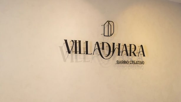 Huoneessa on kaksi puista tuolia ja pyöreä rahi, seinälle kiinnitetty kyltti, jossa lukee ”Villadiana Barrio Creativo”, sekä kattovalaisin.