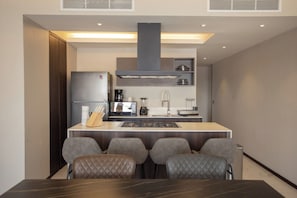 A compact kitchen with a central island, a refrigerator, a stove, and a sink. There are four cushioned chairs around the island.