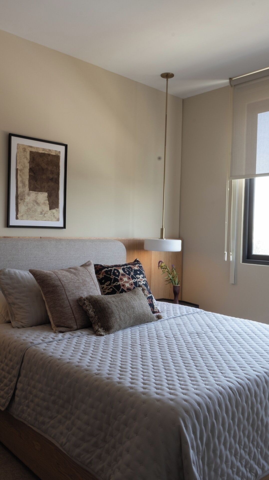 Un dormitorio con cama, almohadas, una obra de arte enmarcada, una lámpara colgante y una ventana.