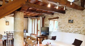 Ein rustikaler Raum mit Steinwänden, Holzbalken, einem weißen Sofa, einem Fernseher und einem Essbereich mit Korbstühlen und einem Tisch.