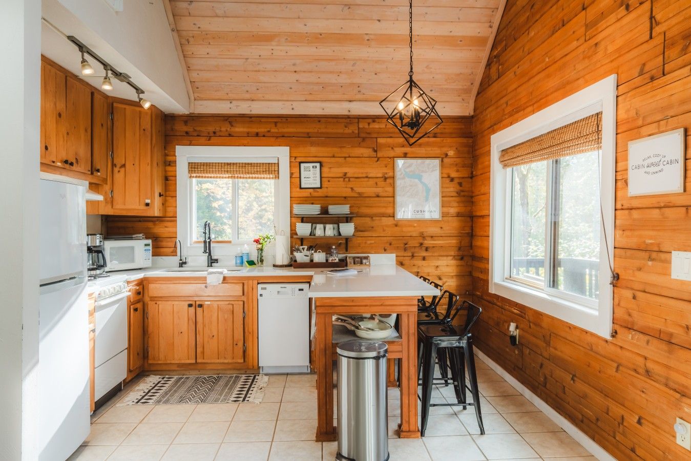L’intérieur d’une cabane avec des murs en bois, une cuisine dotée d’appareils électroménagers blancs, un coin repas avec une table et des chaises, et un luminaire suspendu.