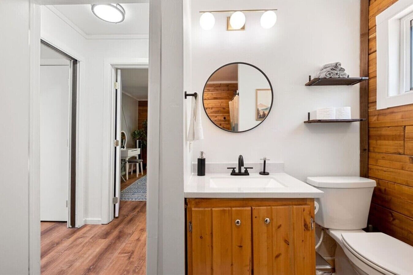 Een badkamer met een houten wastafelmeubel, een ronde spiegel, een witte wasbak en een toilet.