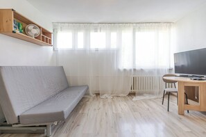 Ein Wohnzimmer mit einer Couch, einem Fernseher, einem Holztisch und einem Fenster mit transparenten Vorhängen.