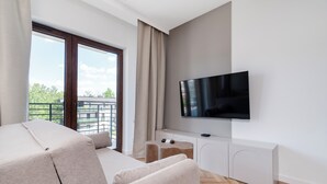 A modern living room with a sofa, a TV mounted on the wall, and a balcony with a view of greenery.