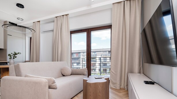 A modern living room with a sofa, a small wooden side table, a TV, and a large window with curtains.