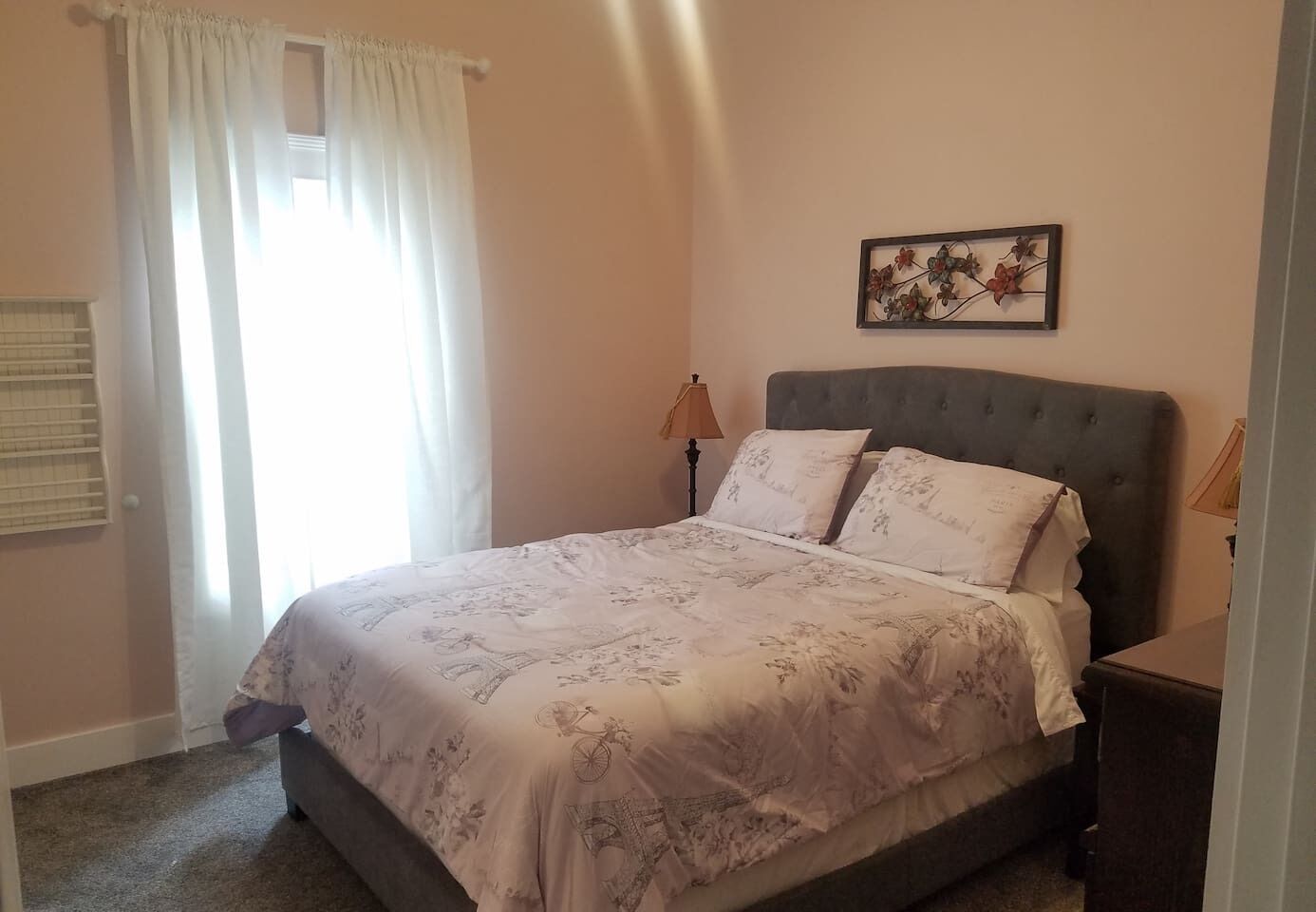 A bedroom with a bed, a nightstand, a lamp, a window with curtains, and a wall-mounted picture.