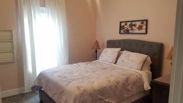 A bedroom with a bed, a nightstand, a lamp, a window with curtains, and a wall-mounted picture.