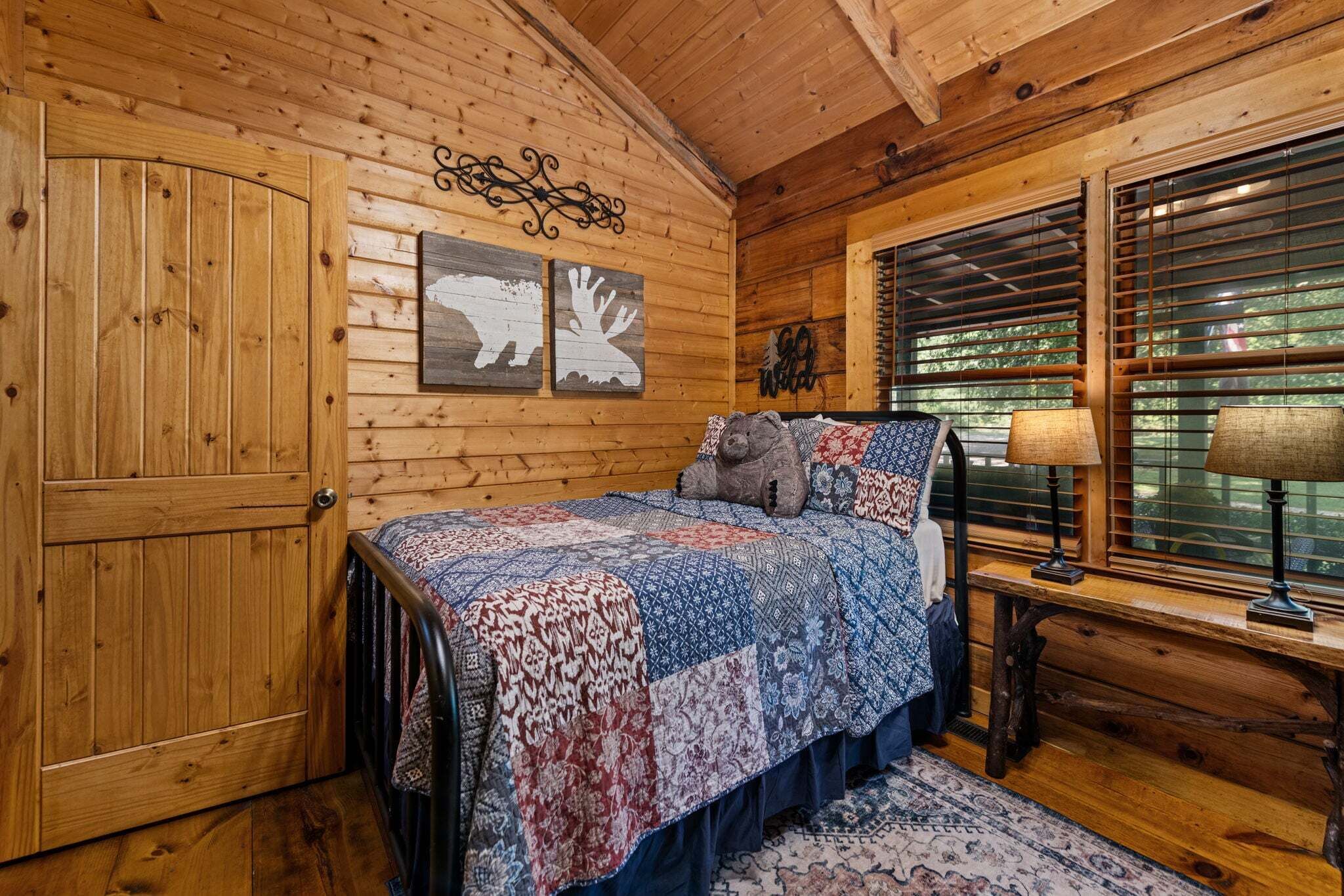 A wooden cabin bedroom with a bed, a window with blinds, and a door.