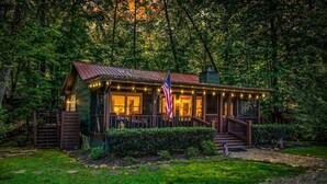 A rustic cabin with a porch, an American flag, and a well-maintained garden. - Creekside Green - Firepit | Gazebo alongside creek | pond (Cherry Log)