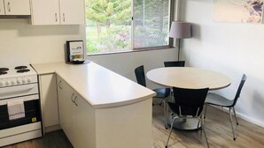 A compact kitchen with a stove, a dining table with four chairs, a window with a view of greenery, and a lamp on the wall. - Apiwa Middleton Beach Front Apartments Albany  (Middleton Beach)