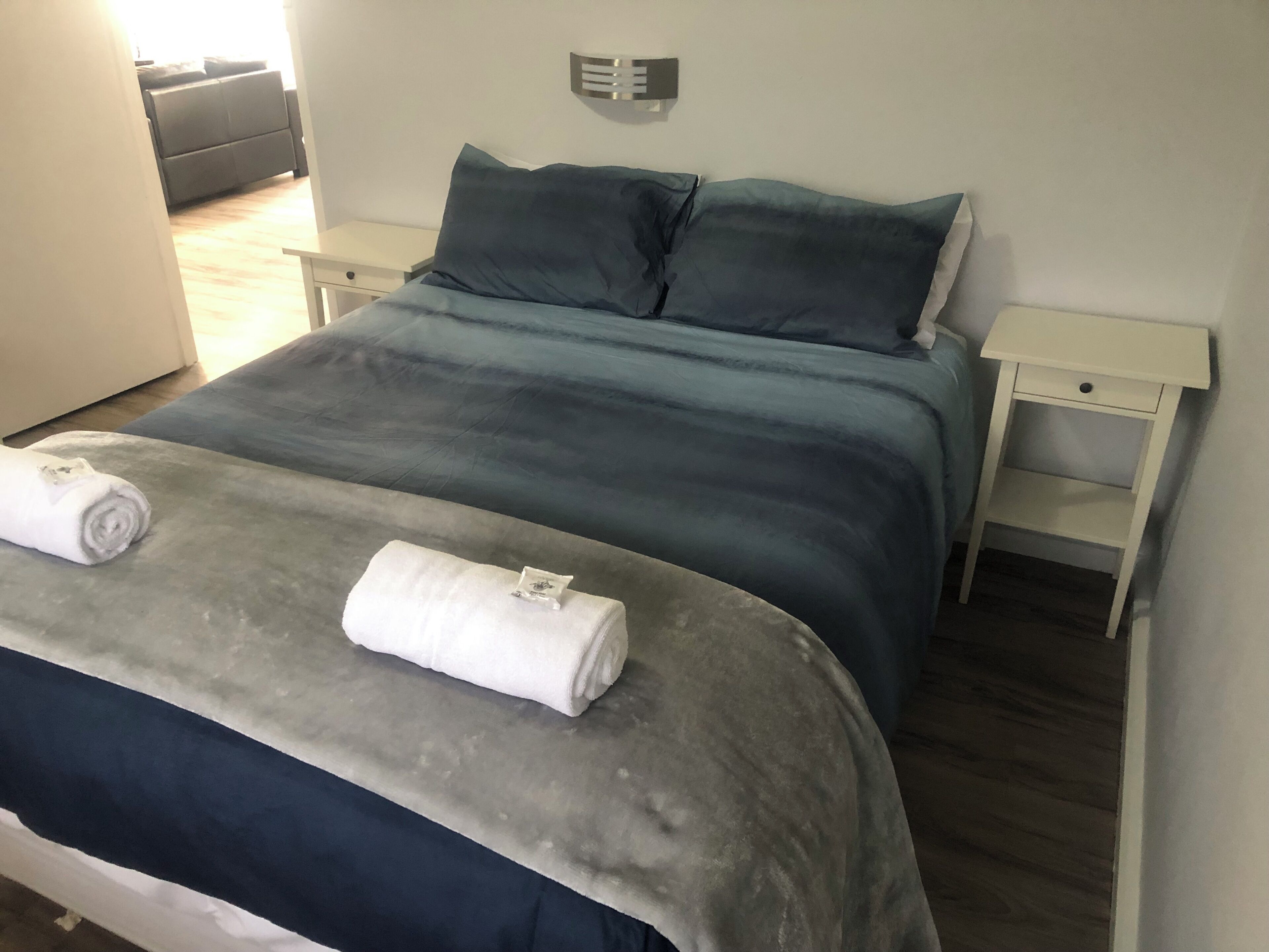 Una cama con colcha gris y azul, dos toallas blancas, una mesita de noche y un aplique de pared.