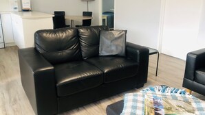 A black leather sofa with a pillow, a small table with a checkered table runner, a wall clock, a lamp, and a framed picture on the wall. - Apiwa Middleton Beach Front Apartments Albany  (Middleton Beach)