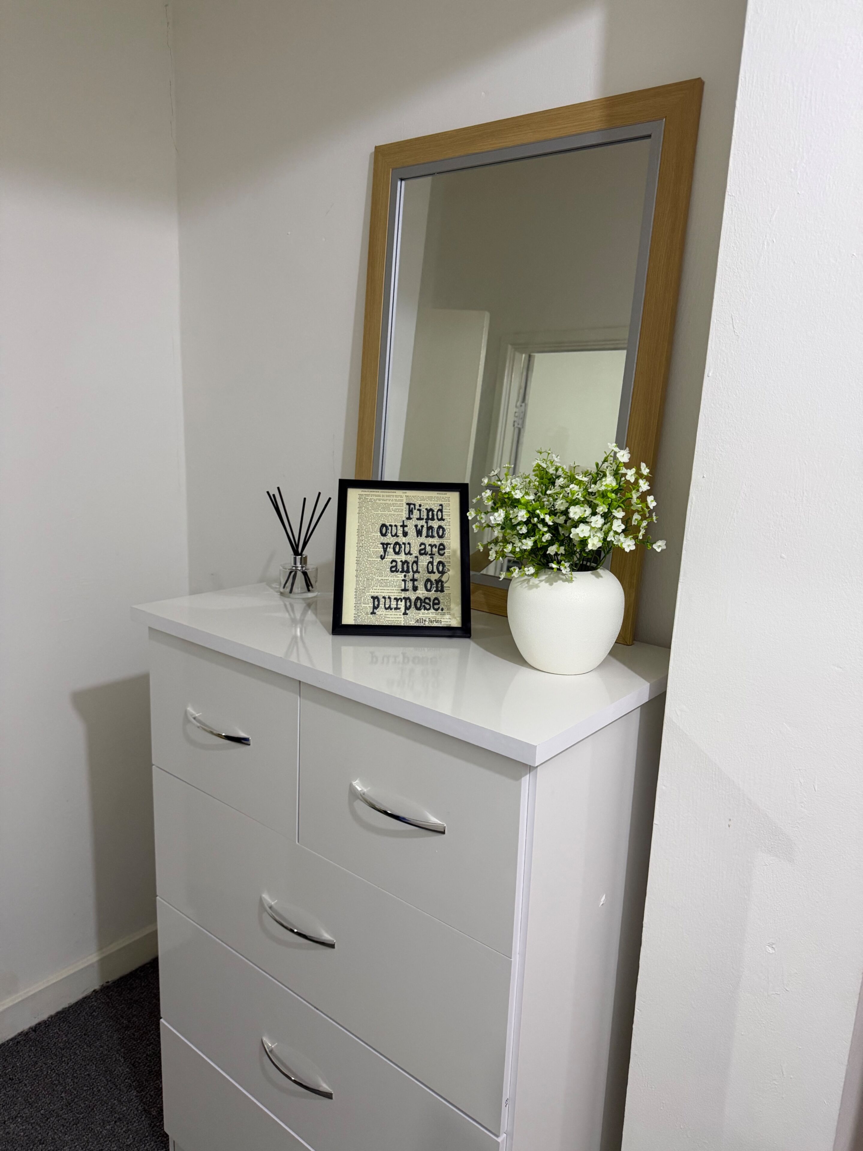 A white dresser with a mirror, a framed inspirational quote, and a vase with white flowers.