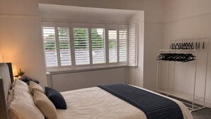 Ein Schlafzimmer mit Bett, Fenster mit Jalousien, Nachttisch mit Lampe und einer Kleiderstange.