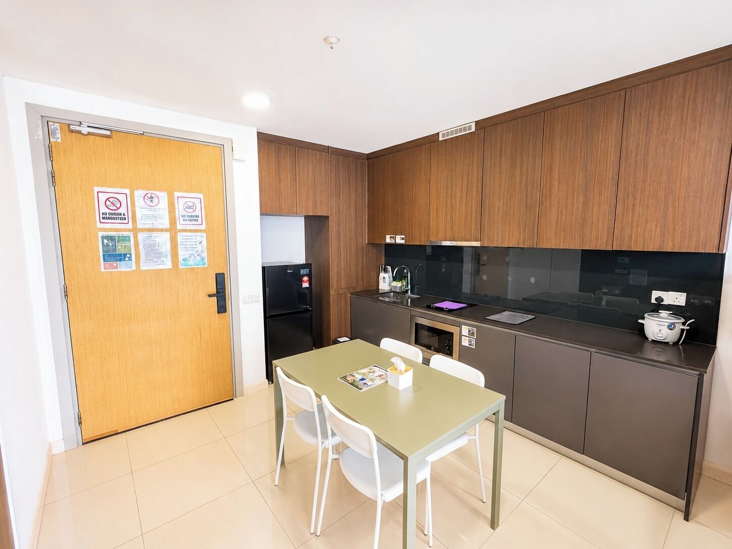 A compact kitchen with a refrigerator, microwave, and stove. There is a dining table with chairs and a wooden door with notices.