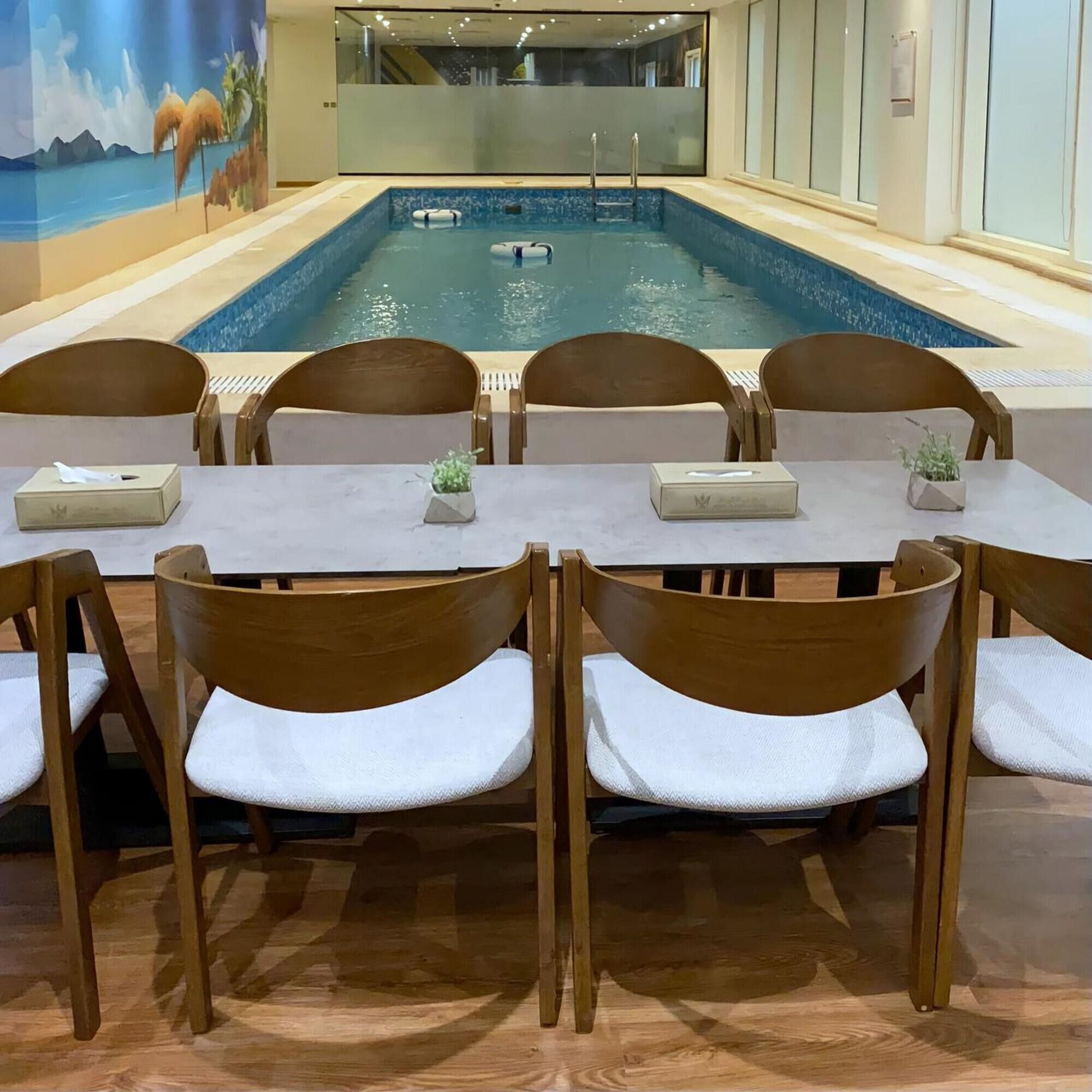 A poolside dining area with wooden chairs and tables, a mural of a beach scene, and a clear view of the pool.
