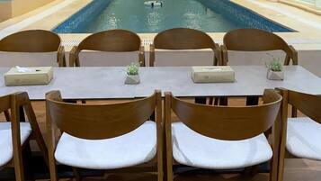 A poolside dining area with wooden chairs and tables, a mural of a beach scene, and a clear view of the pool.