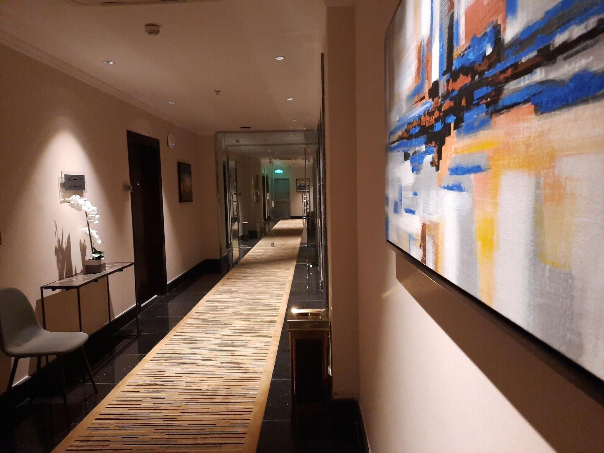 A hotel corridor with a patterned carpet, a large abstract painting on the wall, and a reception desk with a chair.