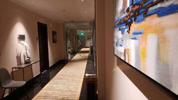 A hotel corridor with a patterned carpet, a large abstract painting on the wall, and a reception desk with a chair.