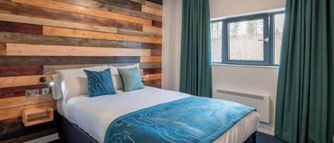 A bedroom with a wooden headboard, a bed with a blue patterned bedspread, a bedside table, and a window with curtains.