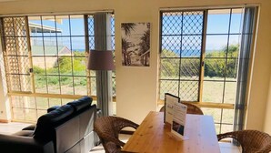 Una habitación con sofá, una mesa de comedor con sillas, una lámpara y ventanas amplias con vistas al exterior.
