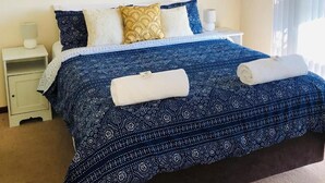 Una cama con un edredón azul y blanco estampado, una mesita de noche blanca con una lámpara y una ventana con cortinas.