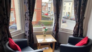 Una habitación con dos sillones negros, una mesita de madera con un jarrón de flores y vistas a la calle a través de amplios ventanales con cortinas.