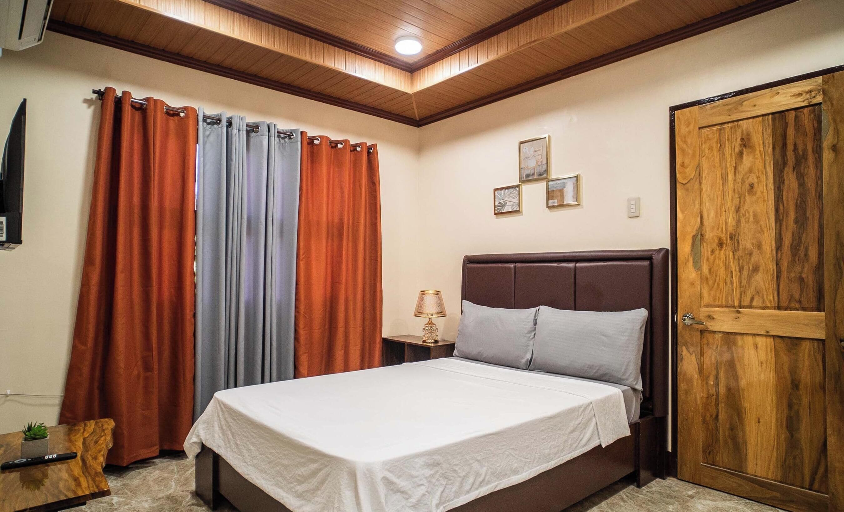 A bedroom with a bed, curtains, a nightstand, a lamp, and a wooden door.