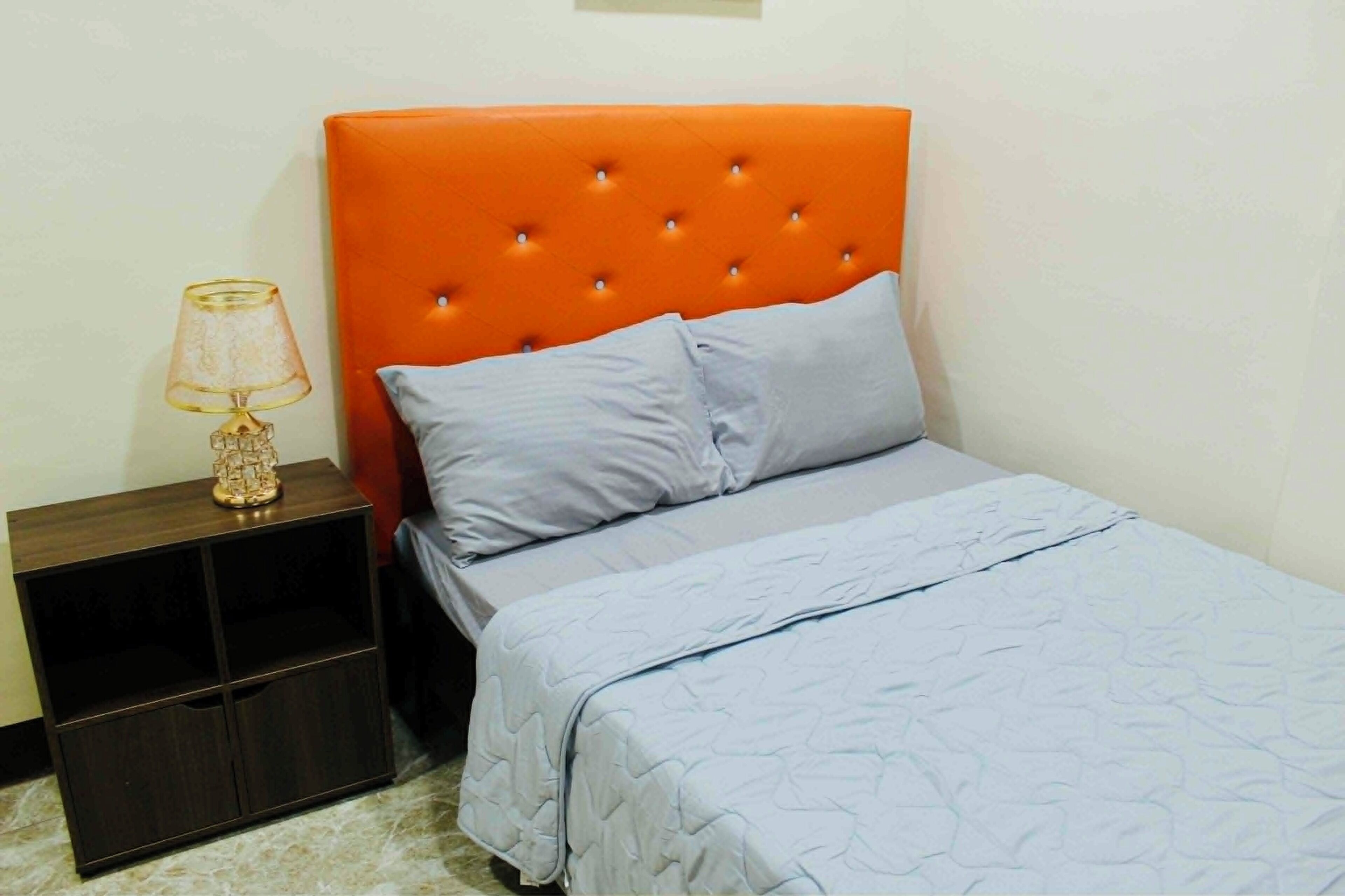 Quarto com cabeceira laranja com textura em botão, uma cama de solteiro com colcha azul claro, mesa de cabeceira com abajur e um armário pequeno.