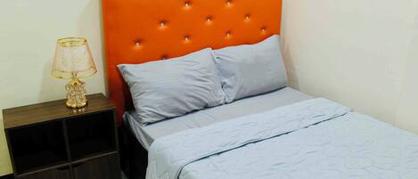 Quarto com cabeceira laranja com textura em botão, uma cama de solteiro com colcha azul claro, mesa de cabeceira com abajur e um armário pequeno.