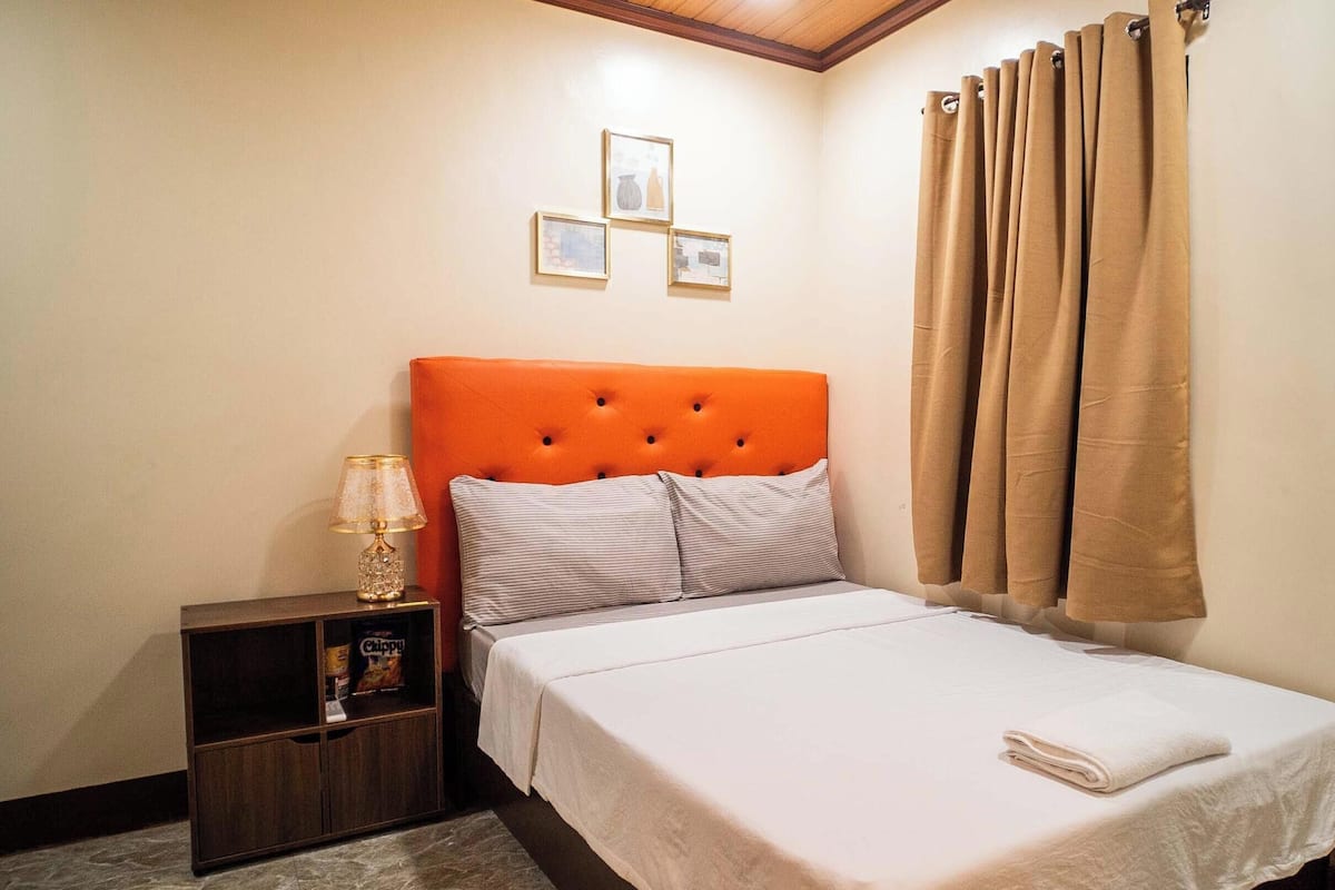 Un dormitorio con un cabecero naranja capitonado, una colcha blanca, una mesita de noche con una lámpara y un estante con aperitivos.