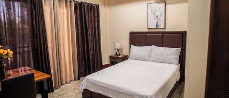 A hotel room with a bed, a desk, a chair, a nightstand, a lamp, and a framed picture on the wall.