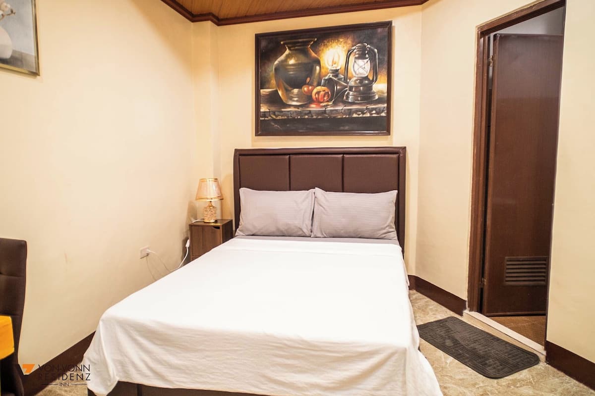 Una habitación de hotel con cama, mesita de noche, lámpara, un cuadro sobre la cama y una puerta.