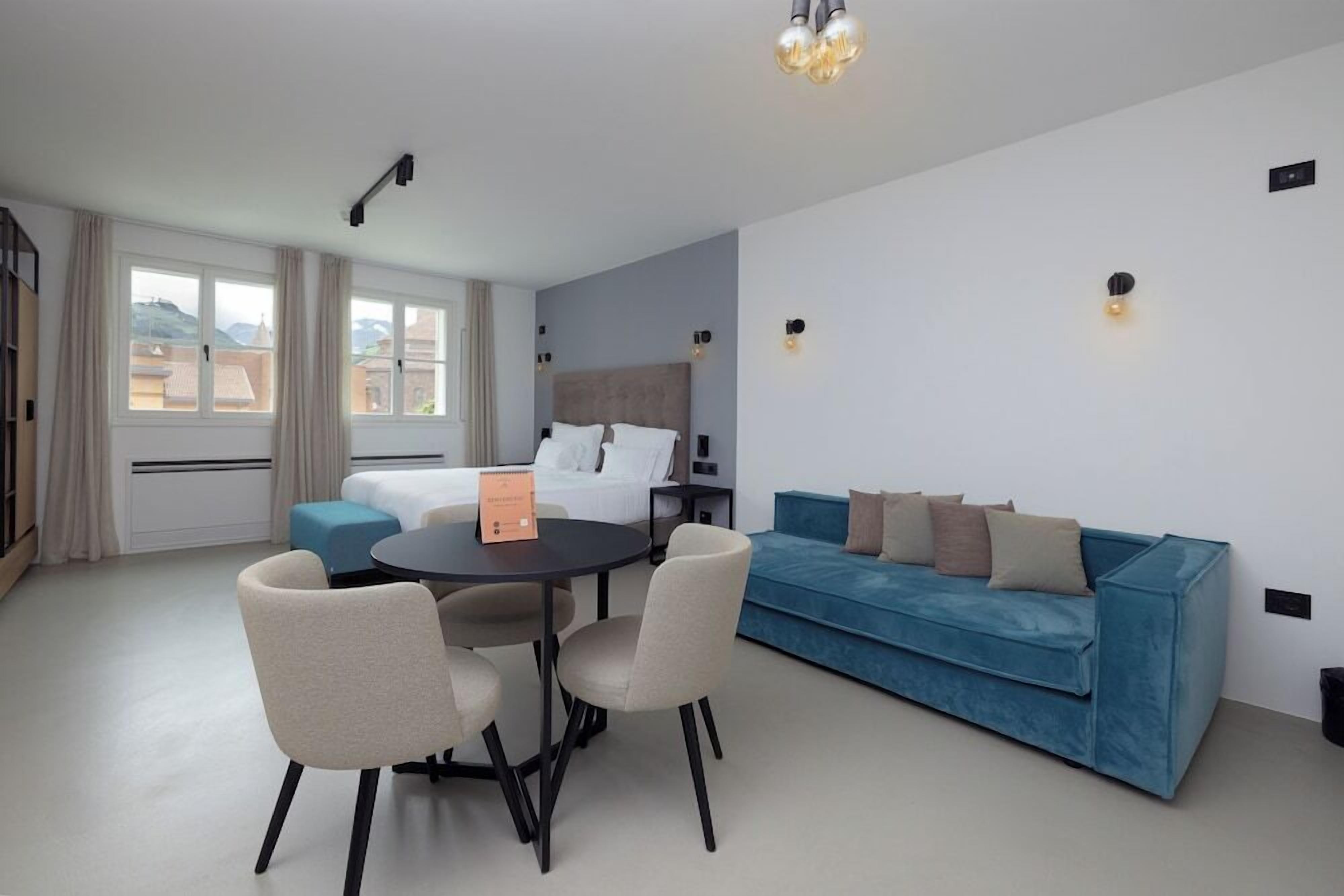 A modern hotel room with a bed, a blue sofa, a dining table with chairs, and a view of the cityscape through large windows.
