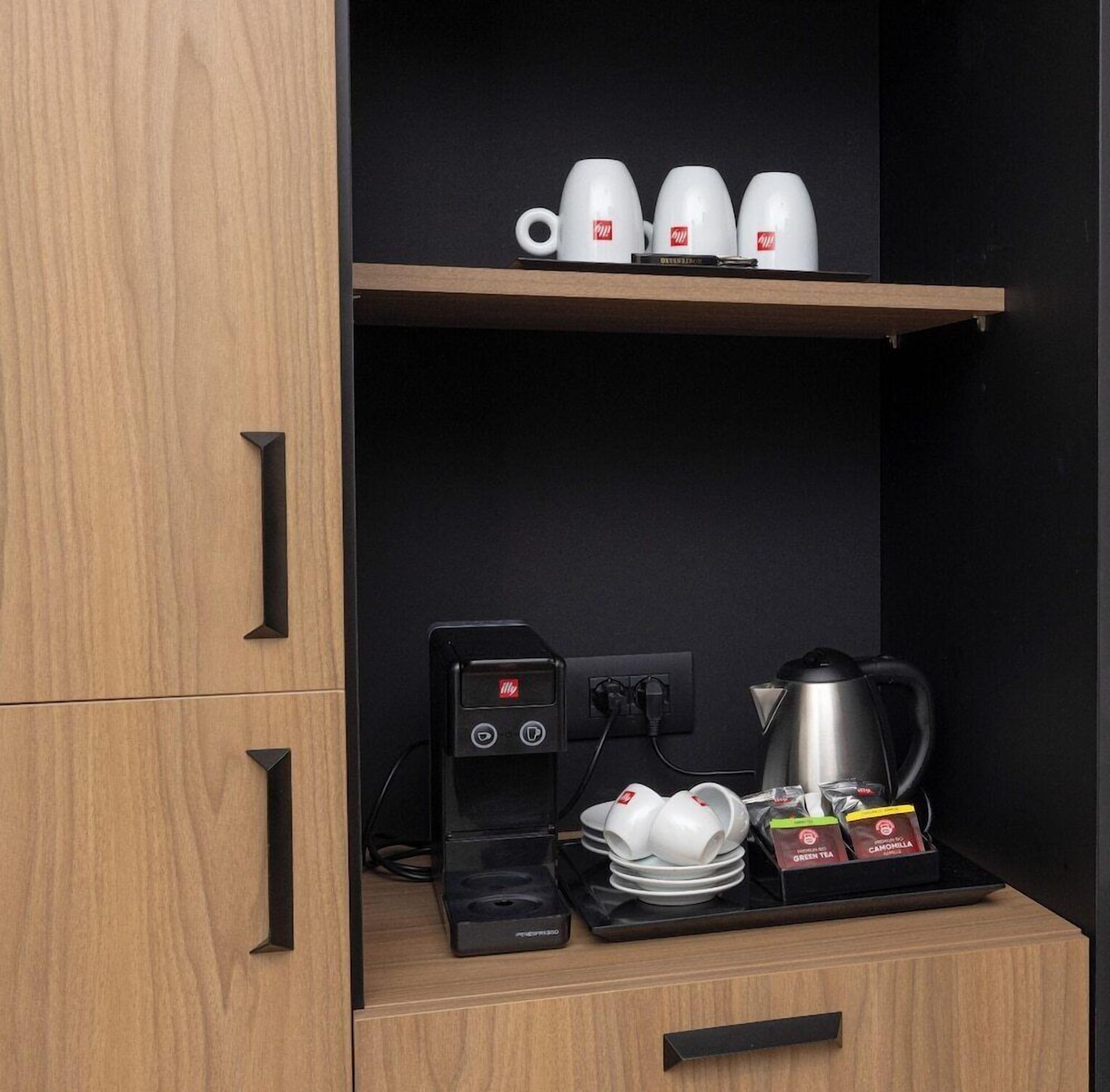 Una kitchenette con cafetera, tetera, una pila de tazas blancas y una caja de té.