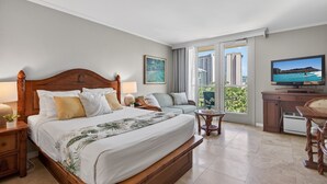 Habitación de hotel con una cama grande, televisión, sofá, una mesita y un balcón con vista a la ciudad.