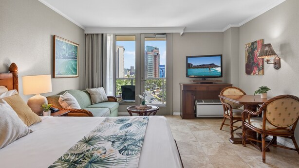 Habitación de hotel con cama, sofá, mesa de comedor con sillas, televisor y balcón con vista a la ciudad.