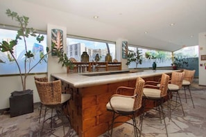 A bar area with a wooden counter and wicker chairs, large windows with city views, and indoor plants. - Island Retreat at the Luana Waikiki (Honolulu)